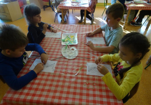 Czwórka dzieci siedzi przy stoliku, wykleja wydzieranymi kawałkami papieru sylwetę Syrenki Warszawskiej.